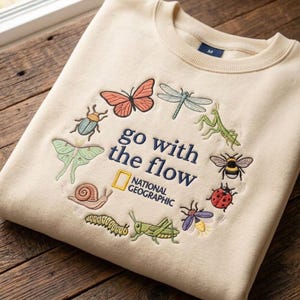 Puede incluir: Una sudadera de cuello redondo color crema con un bordado circular de varios insectos, el texto "go with the flow" y el logotipo de National Geographic en hilo azul oscuro y amarillo en la parte frontal del pecho.