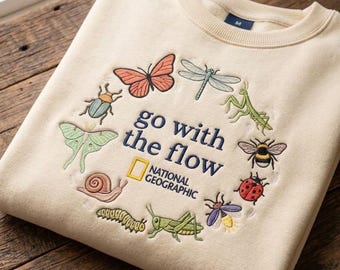 May include: A cream-colored crewneck sweatshirt featuring an embroidered circular arrangement of various insects and the text "go with the flow" alongside the National Geographic logo in the center. The design includes a butterfly, dragonfly, mantis, bee, ladybug, firefly, grasshopper, caterpillar, snail, moth, and beetle.
