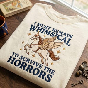 以下が含まれることがあります： クリーム色のクルーネックトレーナー。ダークブルーの刺繍で「I MUST REMAIN WHIMSICAL TO SURVIVE THE HORRORS」という文字が施されています。パーティーハットを被った翼のある馬の刺繍と、その周りに小さな金色の星がデザインされています。