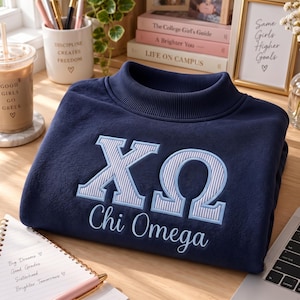 May include: A navy blue sweatshirt featuring a mock neck collar and embroidered Greek letters Chi Omega in light blue with a white striped pattern. The text Chi Omega is stitched in light blue cursive script below the large Greek letters.