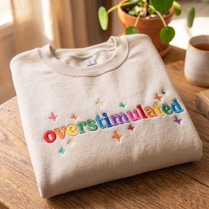 Op de afbeelding: Een beige sweater met ronde hals waarop het woord overstimulated is geborduurd in een levendig regenboogverloop. Het ontwerp is geaccentueerd met kleine, meerkleurige geborduurde sterren die rondom de tekst op de borst zijn verspreid.