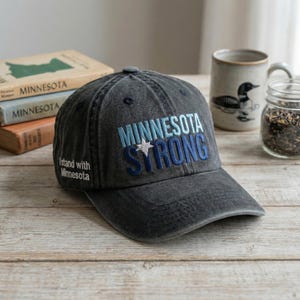 以下が含まれることがあります： 青と白の星が付いた「MINNESOTA STRONG」の文字が刺繍された、チャコールグレーの野球帽。「I stand with Minnesota」というフレーズが側面に刺繍されています。帽子は木製の表面にあります。