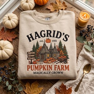 Pode incluir: Uma camisola bege de gola redonda com uma impress&atilde;o gr&aacute;fica de uma casa de pedra rodeada por pinheiros e ab&oacute;boras. O texto diz HAGRID'S est. 1981 PUMPKIN FARM MAGICALLY GROWN, com uma placa a apontar para PUMPKINS, POTIONS, PLANTS.