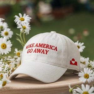 May include: A white baseball cap with the embroidered text "MAKE AMERICA GO AWAY" in red. A small flag is on the side. The cap is surrounded by white daisies.