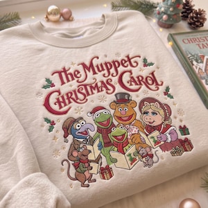 May include: A cream-colored crewneck sweatshirt featuring an embroidered design of The Muppet Christmas Carol characters singing from books. The embroidery includes Gonzo, Kermit, Fozzie Bear, Miss Piggy, and mice, accented with holly and snowflakes.