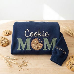 May include: Navy blue sweatshirt with the embroidered words "Cookie MOM" and a cookie graphic. The sleeve has "TROOP 1235" embroidered. Chocolate chip cookies and wheat stalks are scattered around the sweatshirt.