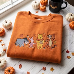 Puede incluir: Una sudadera naranja de cuello redondo con un diseño bordado de Igor, Piglet, Winnie the Pooh sosteniendo un tarro de miel y Tigger. Los personajes están rodeados de pequeñas hojas de otoño y bellotas bordadas en el pecho.