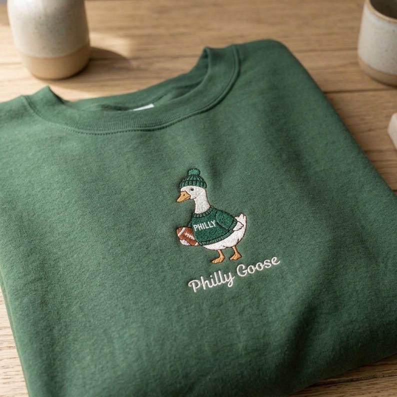 May include: A forest green crewneck sweatshirt featuring an embroidered graphic of a white goose wearing a green knit hat and a green jumper with the word PHILLY printed on it. The goose holds a brown football. The text Philly Goose is below.