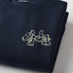 Op de afbeelding: Marineblauw sweatshirt met een crèmekleurige illustratie van vier draken die een spelletje spelen. De draken hebben vleugels en zijn rond een tafel met spelstukken geplaatst. Het sweatshirt is opgevouwen.
