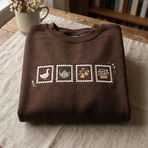 Puede incluir: Una sudadera marrón de cuello redondo con diseños de sellos bordados. Los sellos presentan un ganso, una tetera, peras y una cesta de flores. El bordado es en blanco, verde, amarillo y marrón. La sudadera está doblada sobre una superficie de color neutro.