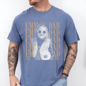 Puede incluir: Camiseta azul con una foto de una ni&ntilde;a con gafas de sol y un chaleco. El texto "EMMA'S DAD" est&aacute; impreso en dorado, junto con "EST. 2011". La camiseta la lleva una persona con tatuajes en el brazo.