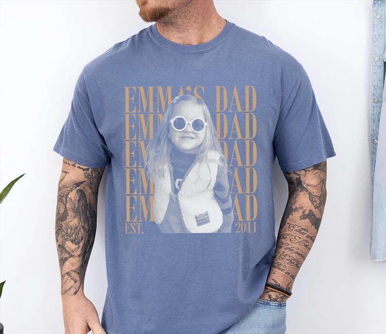 Puede incluir: Camiseta azul con una foto de una ni&ntilde;a con gafas de sol y un chaleco. El texto "EMMA'S DAD" est&aacute; impreso en dorado, junto con "EST. 2011". La camiseta la lleva una persona con tatuajes en el brazo.