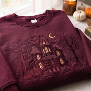 Pode incluir: Uma camisola de gola redonda em bordeaux profundo com um bordado intrincado de uma casa assombrada, árvores sem folhas, abóboras e morcegos sob uma lua crescente. O design é apresentado em tons de dourado e castanho na parte frontal.