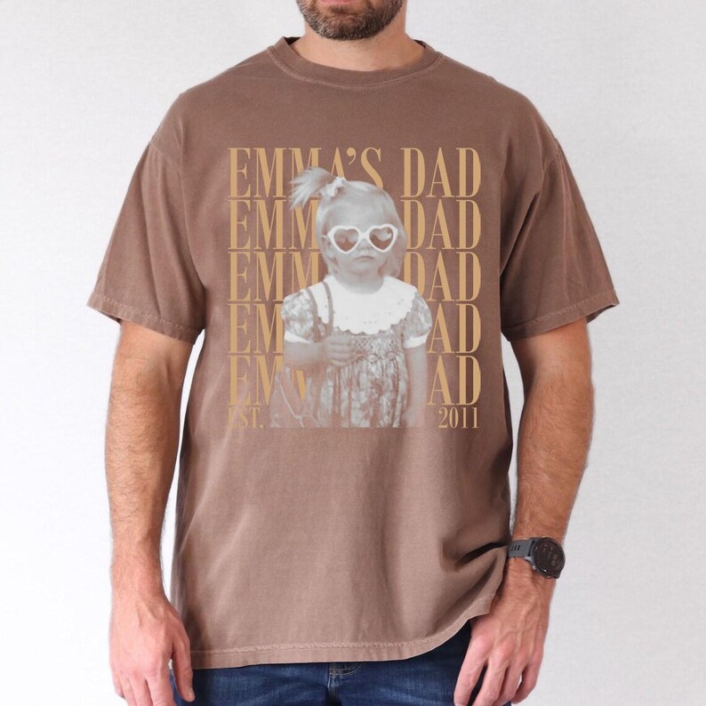 Puede incluir: Camiseta marr&oacute;n con una imagen en sepia de un ni&ntilde;o con gafas de sol en forma de coraz&oacute;n. El texto "EMMA'S DAD" est&aacute; impreso verticalmente a la izquierda y "DAD" a la derecha. El a&ntilde;o "2011" est&aacute; impreso en la parte inferior.