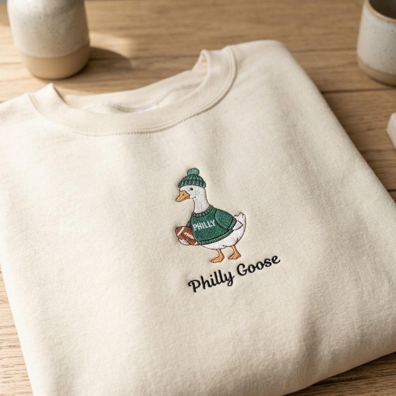 May include: A cream-colored crewneck sweatshirt featuring an embroidered graphic of a white goose wearing a green knit hat and a green sweater with the word PHILLY printed on it, holding a brown football. The text Philly Goose is embroidered below.