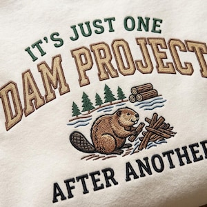 Puede incluir: Una sudadera color crema con un diseño bordado de un castor marrón construyendo una presa con troncos. El texto dice IT'S JUST ONE DAM PROJECT AFTER ANOTHER en letras mayúsculas de color verde oscuro y marrón, junto a pinos verdes.