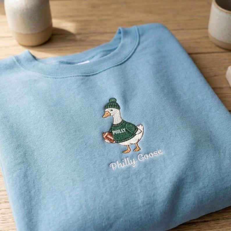 May include: A light blue crewneck sweatshirt featuring an embroidered graphic of a white goose wearing a green knit hat and a green sweater with the word PHILLY printed on it. The goose holds a brown football, and the text Philly Goose is below.