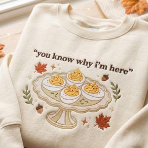May include: A cream-colored crewneck sweatshirt featuring brown embroidered text that reads, "you know why i'm here". Below the text is an embroidered illustration of four deviled eggs on a pedestal platter, surrounded by autumn leaves and acorns.
