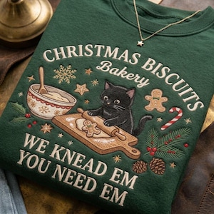 Puede incluir: Una sudadera verde oscuro con texto bordado que dice CHRISTMAS BISCUITS BAKERY WE KNEAD EM YOU NEED EM. El diseño incluye un gato negro usando un rodillo sobre masa, un tazón, un hombre de jengibre y ramas de pino.