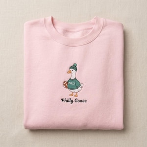 May include: A light pink crewneck sweatshirt featuring an embroidered graphic of a white goose wearing a green knit hat and a green sweater with the word PHILLY printed on it. The goose holds a brown football, and the text Philly Goose is below.