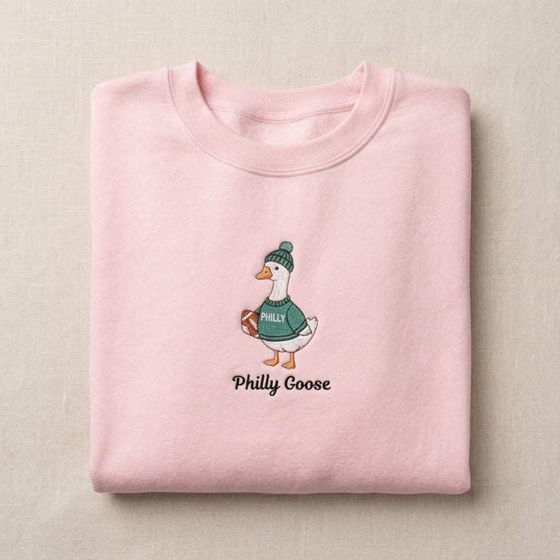 May include: A light pink crewneck sweatshirt featuring an embroidered graphic of a white goose wearing a green knit hat and a green sweater with the word PHILLY printed on it. The goose holds a brown football, and the text Philly Goose is below.