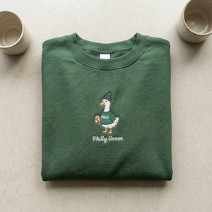 May include: A forest green crewneck sweatshirt featuring an embroidered graphic of a white goose wearing a green beanie and a green sweater with the word PHILLY printed on it. The goose holds a brown football, and the text Philly Goose is below.