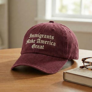 Könnte beinhalten: Burgunderfarbene Baseballkappe mit dem gestickten Text "Immigrants Make America Great" in Weiß. Die Kappe ist aus einem weichen Material gefertigt und hat einen gebogenen Schirm. Die Kappe liegt auf einer Holzoberfläche.