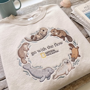 May include: A beige crewneck sweatshirt featuring an embroidered circle of various aquatic animals including otters, a platypus, a manatee, and a seal. The text "go with the flow" and the "NATIONAL GEOGRAPHIC" logo are embroidered in the centre.
