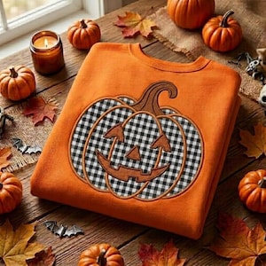 Op de afbeelding: Een oranje sweater met ronde hals en een groot geborduurd pompoenontwerp. De pompoen heeft een zwart-wit geblokt vichy-patroon met een bruine omlijning en een uitgesneden jack-o'-lantern gezicht met driehoekige ogen en een gekartelde mond.