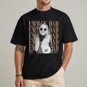 Puede incluir: Camiseta negra con una imagen en tonos sepia de un ni&ntilde;o con gafas de sol y un chaleco. El texto "EMMA'S DAD" est&aacute; impreso verticalmente en un patr&oacute;n repetido, con "EST. 2011" en la parte inferior.