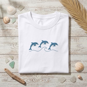 May include: A white short-sleeved t-shirt featuring an embroidered design of three blue dolphins leaping above a single blue wave line. The embroidery is centered on the chest area of the garment, which has a classic crew neckline and a plain white finish.