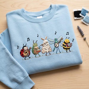 May include: A light blue crewneck sweatshirt featuring an embroidered design of five insects playing musical instruments. The insects include a ladybird with a guitar, a beetle with a drum, a moth with a flute, a dragonfly with a banjo, and a bee with an accordion, all accompanied by black musical note symbols.