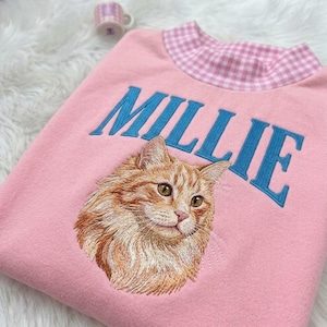 Puede incluir: Una sudadera rosa claro con cuello alto de patrón de cuadros vichy rosa y blanco. En la parte frontal aparece el nombre MILLIE bordado en letras azules sobre un bordado detallado y realista de un gato atigrado de color naranja.