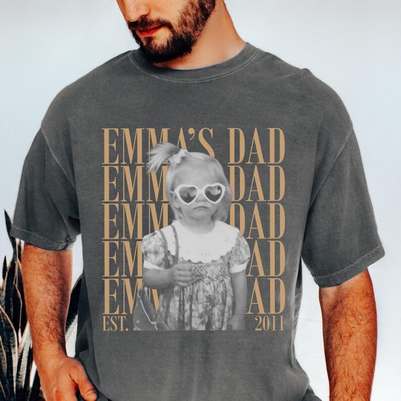 Puede incluir: Camiseta gris oscuro con una imagen en blanco y negro de una ni&ntilde;a peque&ntilde;a con gafas de sol en forma de coraz&oacute;n. El texto "EMMA'S DAD" est&aacute; impreso en un patr&oacute;n repetido en color marr&oacute;n claro. La camiseta tambi&eacute;n incluye el texto "EST. 2011".