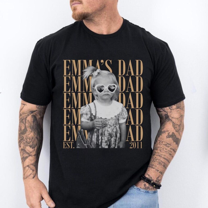 Puede incluir: Camiseta negra con una imagen en sepia de una ni&ntilde;a peque&ntilde;a con gafas de sol en forma de coraz&oacute;n. El texto "EMMA'S DAD" se repite verticalmente en dorado, con "EST. 2011" debajo.