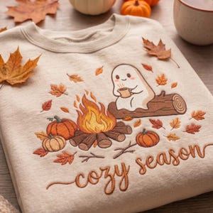 Puede incluir: Una sudadera beige con un diseño bordado de un fantasma blanco sentado en un tronco, sosteniendo una taza junto a una fogata. La escena incluye calabazas naranjas y hojas de otoño, con el texto "cozy season" bordado en la parte inferior.