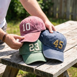 Puede incluir: Tres gorras de béisbol apiladas en rojo, verde y azul sobre una superficie de madera. Cada gorra tiene un número diferente en un color contrastante: 10, 23 y 45. Las gorras tienen un aspecto vintage y desgastado.