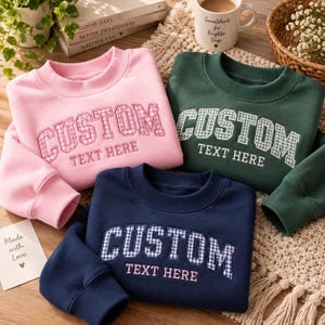 Op de afbeelding: Drie sweaters met ronde hals in roze, donkergroen en marineblauw. Elk heeft geborduurde tekst met CUSTOM in een gingham-patroon en TEXT HERE in een eenvoudig lettertype. De sweaters hebben geribbelde kragen en boorden met een uniform ontwerp.