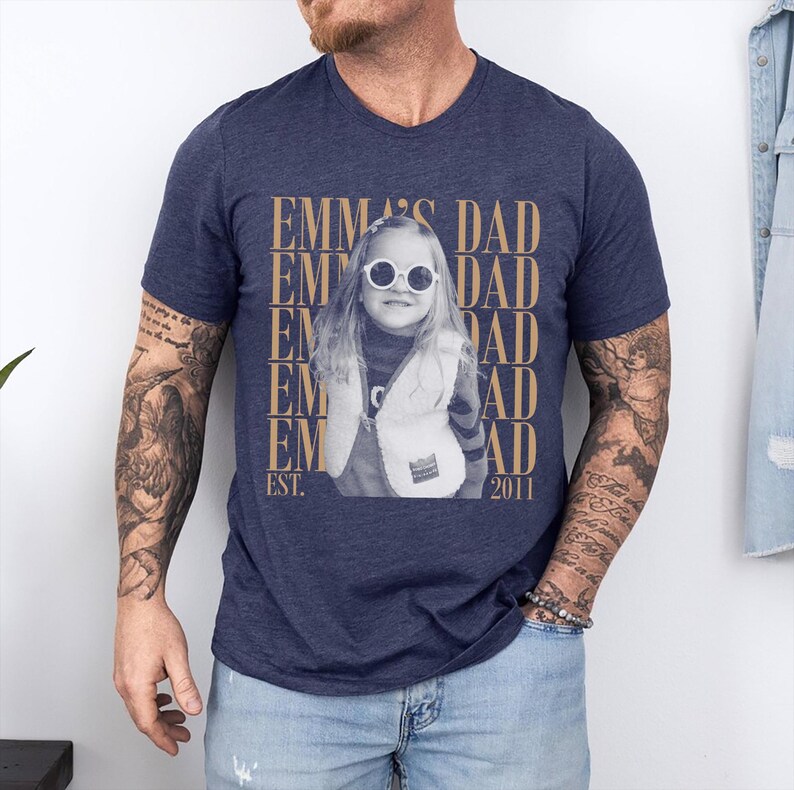 Puede incluir: Camiseta azul marino con cuello en V con una imagen en sepia de un ni&ntilde;o con gafas de sol y un chaleco. El texto "EMMA'S DAD" est&aacute; impreso verticalmente a ambos lados de la imagen, con "EST. 2011" debajo.