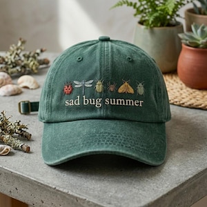 May include: A green baseball cap with the embroidered text "sad bug summer" and various bug illustrations. The cap has an adjustable strap and a curved brim. The cap is made of a soft, textured fabric.