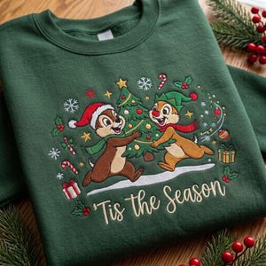May include: A dark green crewneck sweatshirt featuring an embroidered design of two chipmunks decorating a Christmas tree. The scene includes snowflakes, candy canes, and gift boxes, with the text 'Tis the Season' stitched in cream thread.