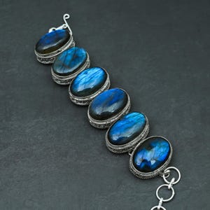 May include: A silver bracelet featuring seven oval-shaped blue labradorite stones set in silver bezels. The stones are surrounded by intricate silver designs.