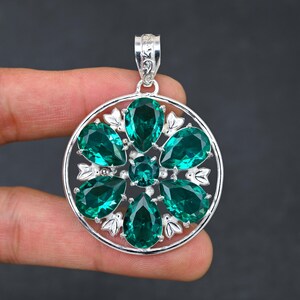 以下が含まれることがあります： 緑色の宝石の花がついた丸いデザインのシルバーペンダント。ペンダントは繊細なフィリグリーパターンで、どんな服装にもエレガントさを加えるのに最適です。