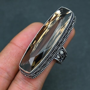 Op de afbeelding: Een rechthoekige ring met rookkwarts edelsteen en een zilveren band. De edelsteen heeft een lichtbruine kleur en is gefacetteerd om het licht te vangen. De zilveren band heeft een sierlijk, gedetailleerd ontwerp.
