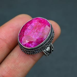 May include: A silver ring with a large, oval-shaped, pink ruby gemstone. The ring has an intricate, ornate design.
