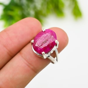 May include: A silver ring featuring a large, oval-cut ruby gemstone. The ruby is a vibrant shade of pink-red, and the ring has a simple, elegant design. The gemstone is held in place by four prongs.