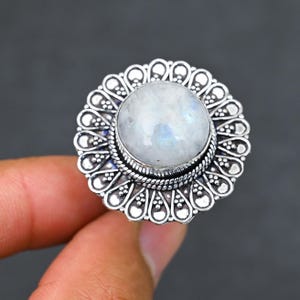 Puede incluir: Anillo plateado con una gran piedra lunar redonda e iridiscente. La piedra lunar está engastada en un bisel detallado de inspiración floral. El anillo tiene un diseño intrincado y texturizado, con un estilo de inspiración vintage.