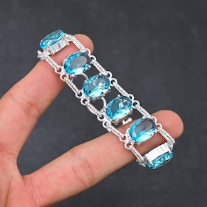 Puede incluir: Pulsera de plata con piedras preciosas ovales de color azul claro. La pulsera tiene un diseño de eslabones rectangulares, con las piedras engastadas en el centro de cada eslabón. La pulsera se muestra sobre un fondo oscuro.