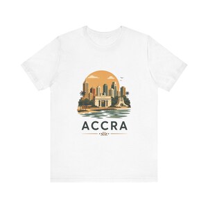 Könnte beinhalten: Ein weißes Kurzarm-T-Shirt mit einer grafischen Illustration der Skyline von Accra, Palmen, einer großen orangefarbenen Sonne und einem Gewässer. Das Wort ACCRA ist in fetten schwarzen Großbuchstaben unter der Stadtszene gedruckt.