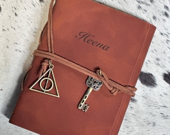 May include: A brown leather journal with a key and a triangle symbol charm attached to the cover. The journal is personalized with the name "Heena".